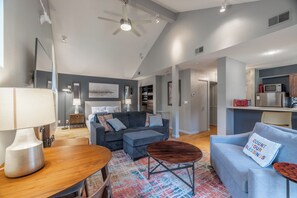 A modern living room with a sofa, a coffee table, and a dining table. There is a kitchen area with a microwave and a refrigerator.