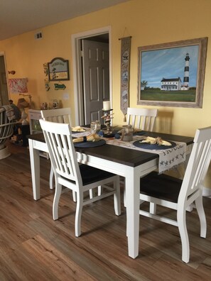 Un coin repas avec une table et des chaises blanches, un tableau représentant un phare et une porte donnant sur une autre pièce.