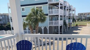 Une terrasse en bois avec deux chaises Adirondack bleues et une petite table, donnant sur une zone riveraine bordée de bâtiments et de palmiers.