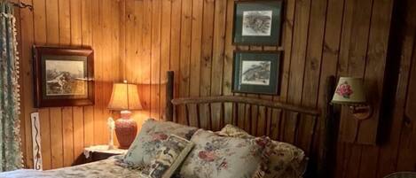 A rustic bedroom with wooden walls, a bed with floral bedding, a ceiling fan, and a wicker chair.