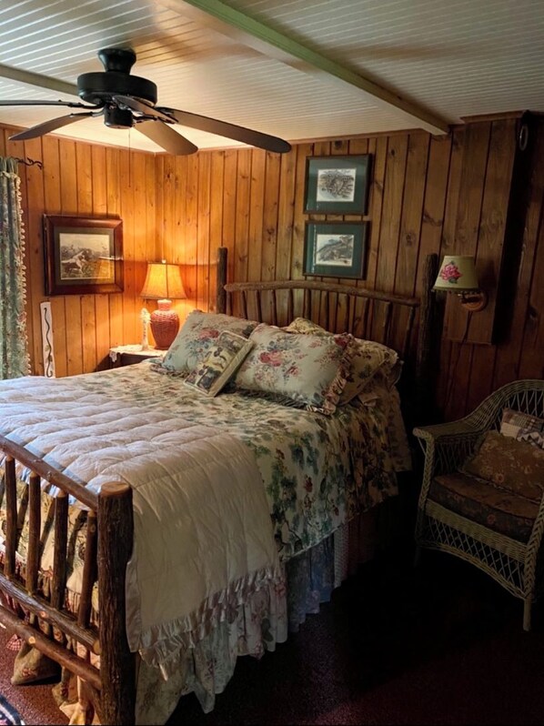 Een rustieke slaapkamer met houten wanden, een bed met bloemenbeddelsels, een plafondventilator en een rieten stoel.