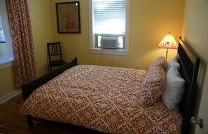 A bedroom with a patterned bedspread, a bedside lamp, a chair, and a window with an air conditioner.