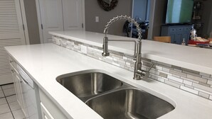 A modern kitchen with a white countertop, two stainless steel sinks, and a backsplash with white and gray tiles. - 4BR Oceanfront Condo in Myrtle Beach  (North Myrtle Beach)