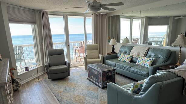 A spacious living room with a large window overlooking the ocean, a ceiling fan, and a leather sofa with colorful cushions. - 4BR Oceanfront Condo in Myrtle Beach  (North Myrtle Beach)