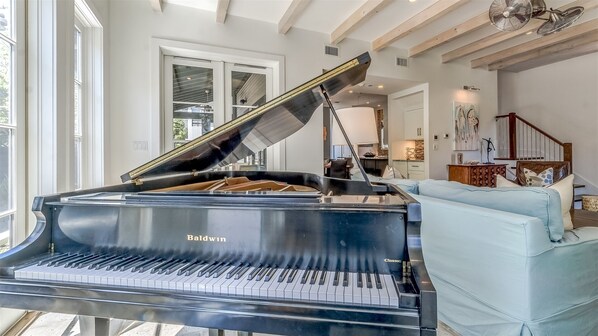 Un piano de cola en un amplio salón con un sofá azul claro y una escalera que sube a un nivel superior.