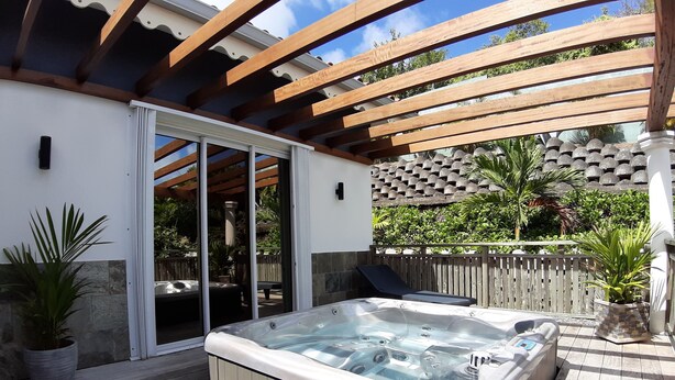 A hot tub under a wooden pergola with a view of a stone wall and greenery.