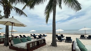A beachside lounge area with wooden loungers, white cushions, and palm trees. - Beachfront condo with full sea view and the facilities of a 5-star resort (Petchaburi)
