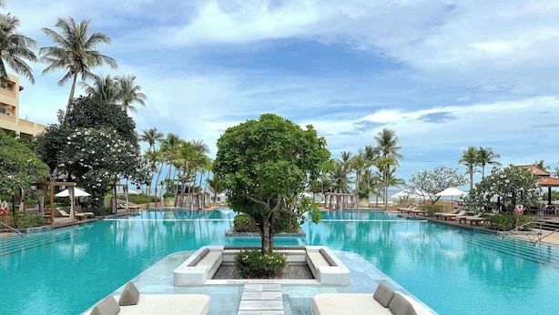 A large swimming pool with lounge areas, surrounded by palm trees and a clear sky. - Beachfront condo with full sea view and the facilities of a 5-star resort (Petchaburi)