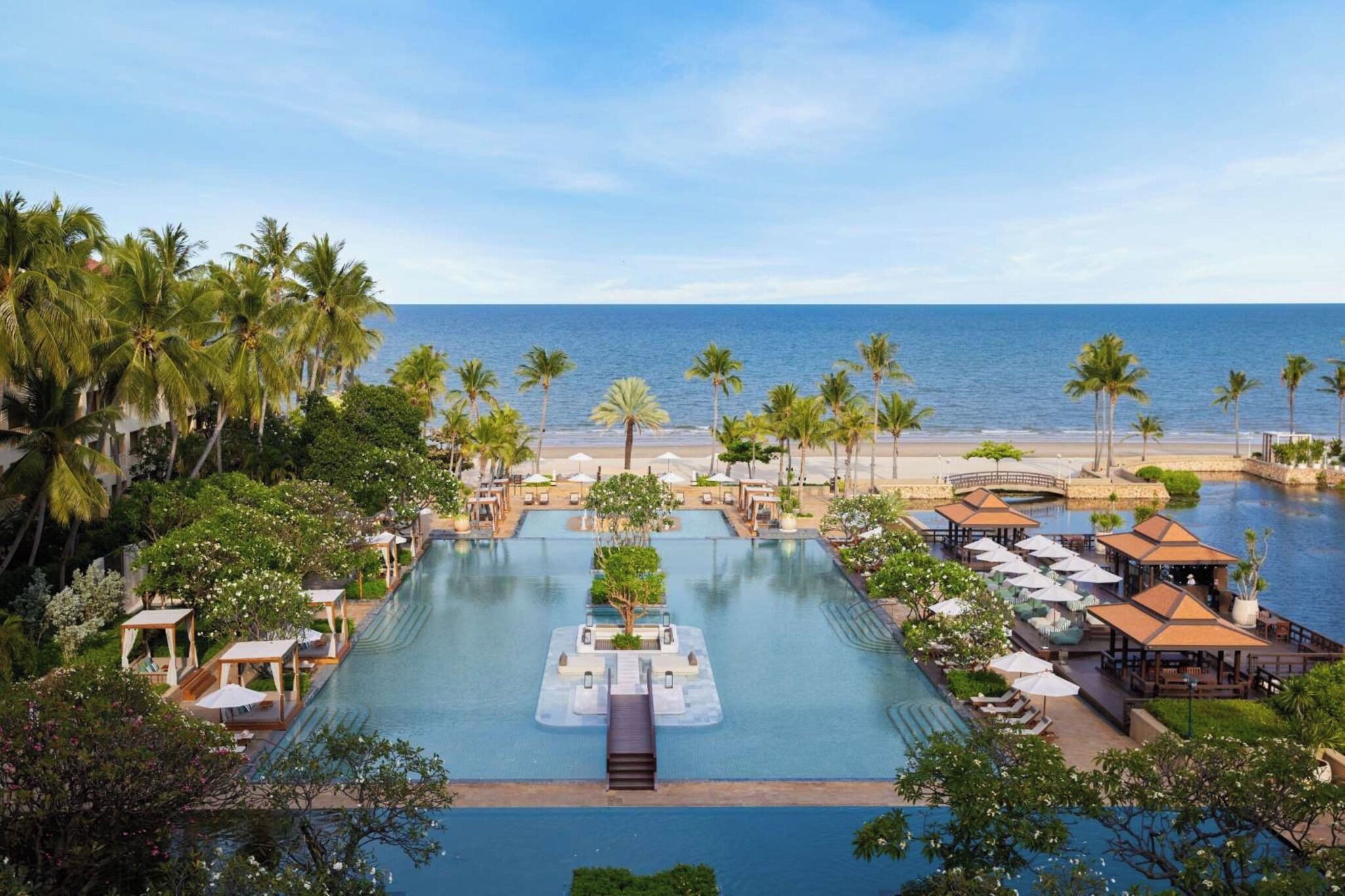 Piscina de resort con un puente, palmeras y vistas a la playa.