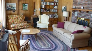 A cozy living room with wooden paneling, a bookshelf, a fireplace, and a floral sofa. - Ocean front house with two private beaches-- on a peninsula--spectacular views! (Milbridge)