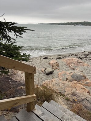 Eine Holztreppe führt zu einem felsigen Strand mit Blick auf das Meer und das entfernte Land.