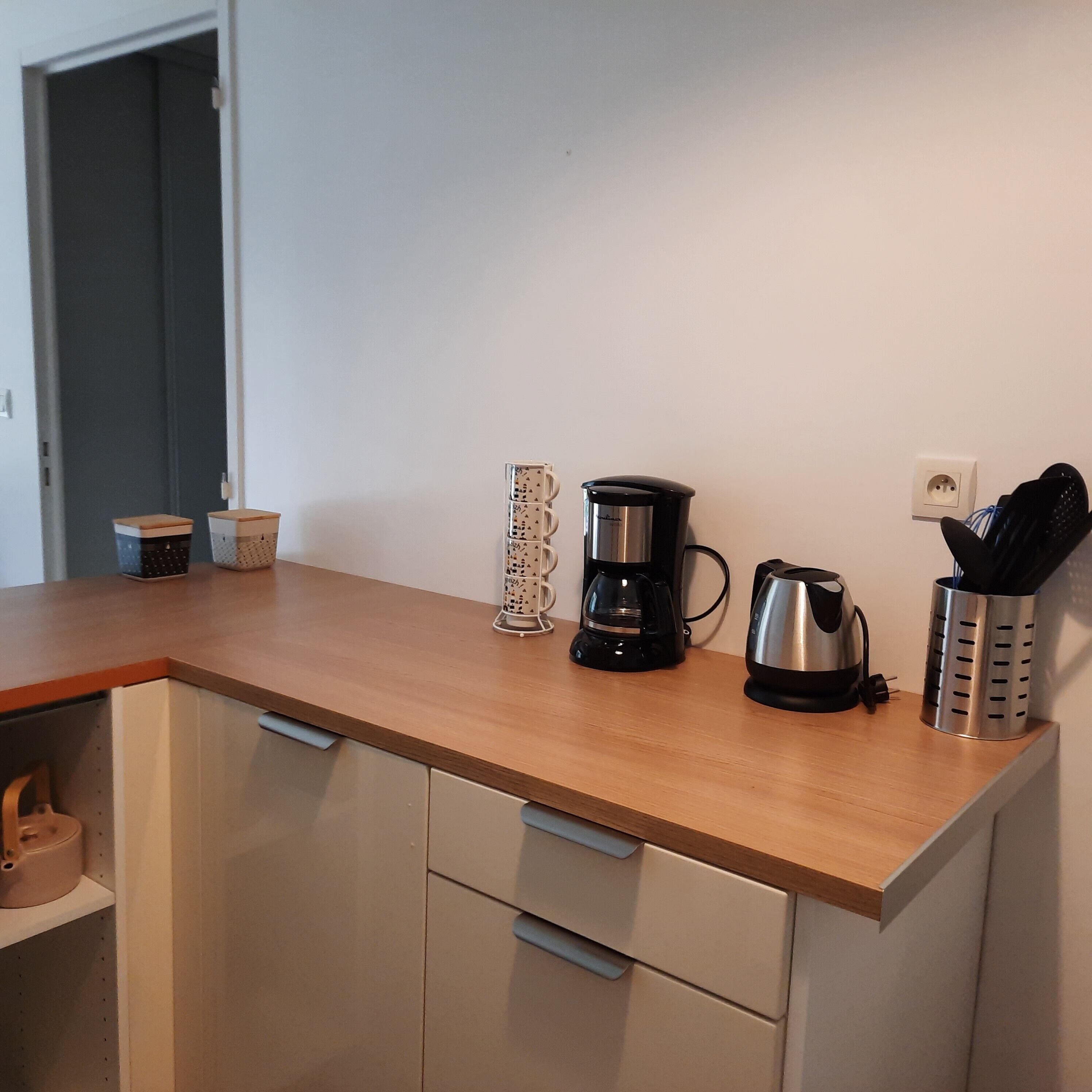 Een keukenblad met een koffiezetapparaat, een waterkoker en een bestekhouder. Daaronder bevinden zich keukenkastjes en een deur naar een andere kamer.