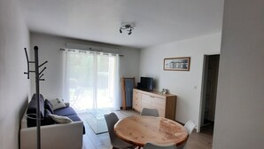 A modern living room with a sofa, a dining table with chairs, a coat rack, and a view of the outdoors through large windows.