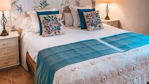 Een slaapkamer met een houten plafond, een bed met bloemenkussens, twee wandlampen en ingelijste kunstwerken.