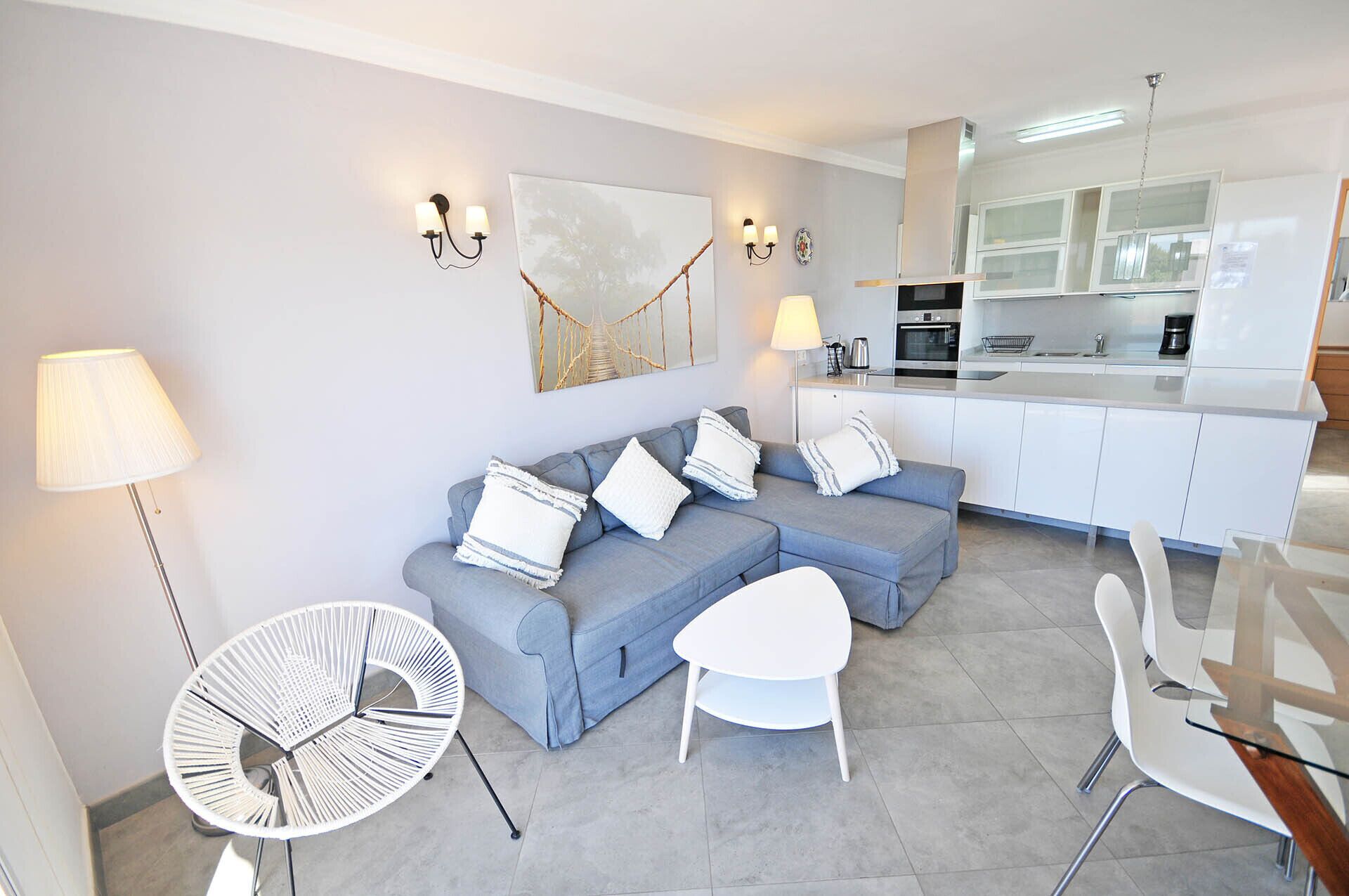 A modern living room with a grey sofa, white and striped cushions, a white coffee table, and a white chair. There is a round white table with a metal frame and a floor lamp. The room has a kitchen area with white cabinets and a stainless steel oven.