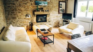 Ein gemütliches Wohnzimmer mit Steinwänden, einem Kamin und einem Fernseher. Zwei weiße Sofas und ein Couchtisch mit Fernbedienung sind vorhanden.