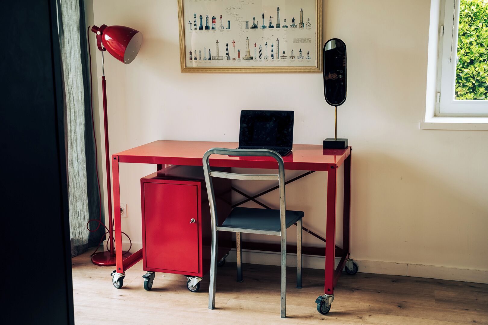 A red desk with a laptop, a red filing cabinet, a metal chair, a red desk lamp, and a framed map on the wall.