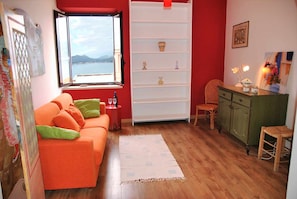 Ein gemütlicher Wohnbereich mit Sofa, einem kleinen Tisch mit Flasche und Glas, einem Bücherregal, einem Sessel und Meerblick durch ein Fenster.