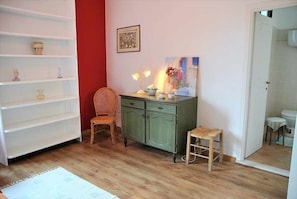 Ein Zimmer mit Holzboden, einem grünen Schrank, einem Korbstuhl, einem kleinen Hocker und einem weißen Regal.
