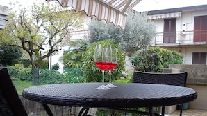 A round outdoor table with a glass of red wine, surrounded by wicker chairs, set on a tiled patio with a view of a garden and a building in the background. - Pretty Casa Margherita, Lugana di Sirmione for your relaxation (Sirmione)