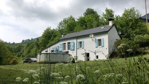 A white house with blue shutters, surrounded by greenery and a car in the foreground.