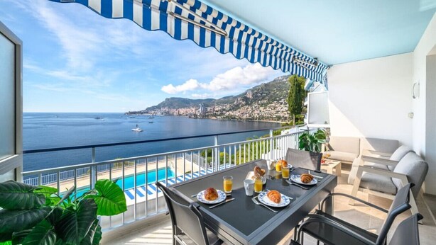 A balcony with a view of the sea, a pool, and a dining table set with plates and glasses.