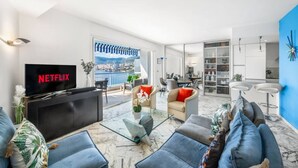 A modern living room with a large flat-screen TV, comfortable seating, a glass coffee table, and a view of a cityscape through large windows.