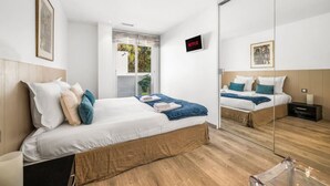 A modern bedroom with a large bed, a bedside table, a mirror, and a window with a view of greenery.
