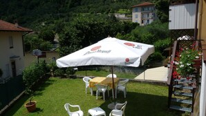 An outdoor seating area with a table and chairs under a large umbrella, surrounded by a well-maintained lawn and a view of a hillside. - TOWNHOUSE WITH GARDEN   (Gavazzo  Di Tenno)
