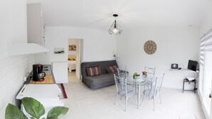 Una cocina moderna con armarios blancos, un comedor con mesa de vidrio y sillas de metal, un sofá gris y una lámpara en el techo.