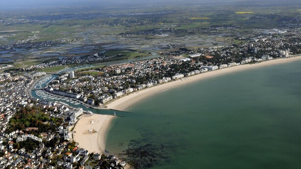 Vista aérea de un pueblo costero con un puerto deportivo, playa de arena y zonas residenciales.