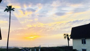 A sunset view from a balcony overlooking a beach with palm trees and a building with outdoor seating.