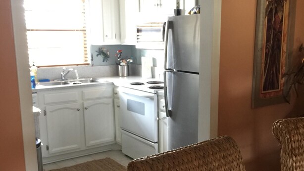 Una cocina con armarios blancos, un refrigerador de acero inoxidable y un ventanal con persianas.
