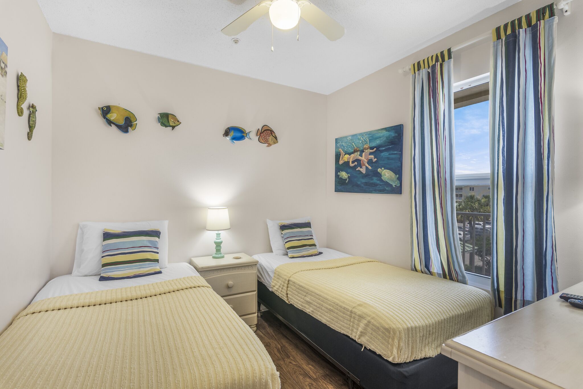 Une chambre avec deux lits, un ventilateur de plafond, un tableau représentant des poissons et une fenêtre avec des rideaux.