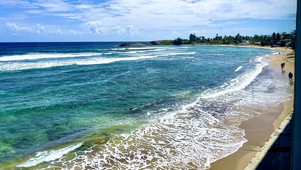 A beach with clear turquoise waters, a sandy shore, and a distant view of a small island with vegetation and buildings. - Villa Caribe by Casa Caribe Vacation Rentals (Aguadilla)