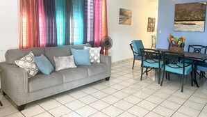 A living room with a grey sofa, blue and white cushions, a dining table with blue chairs, a fan, and colorful curtains. - Villa Caribe by Casa Caribe Vacation Rentals (Aguadilla)