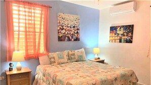 A bedroom with a bed, two bedside tables, a nightstand, a window with curtains, and wall art. - Villa Caribe by Casa Caribe Vacation Rentals (Aguadilla)