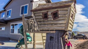 Una casita de juegos de madera con un tobogán, una casa al fondo y un niño cerca.