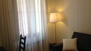 A room with a dark-colored sofa, a wooden chair, a lamp, and a window with sheer curtains.