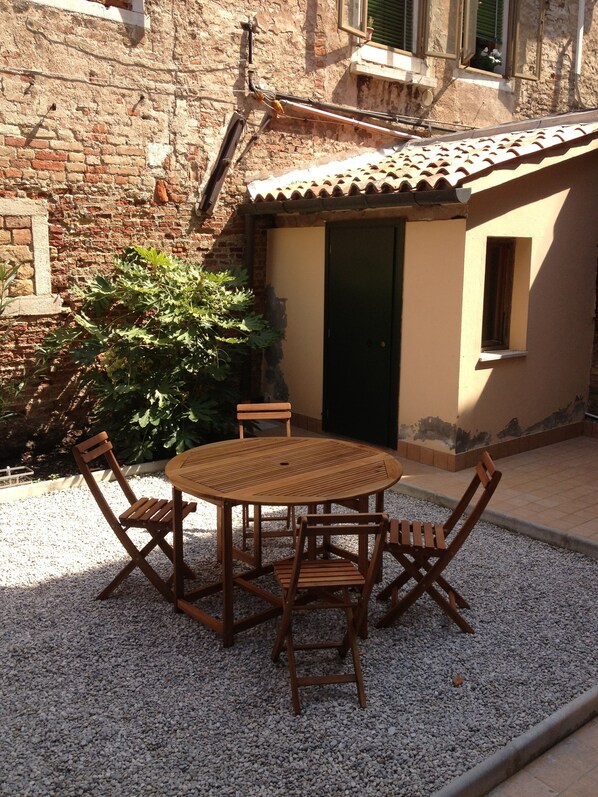 A round wooden table with four chairs in a courtyard with a gravel floor, surrounded by a rustic building with a tiled roof and a green door.