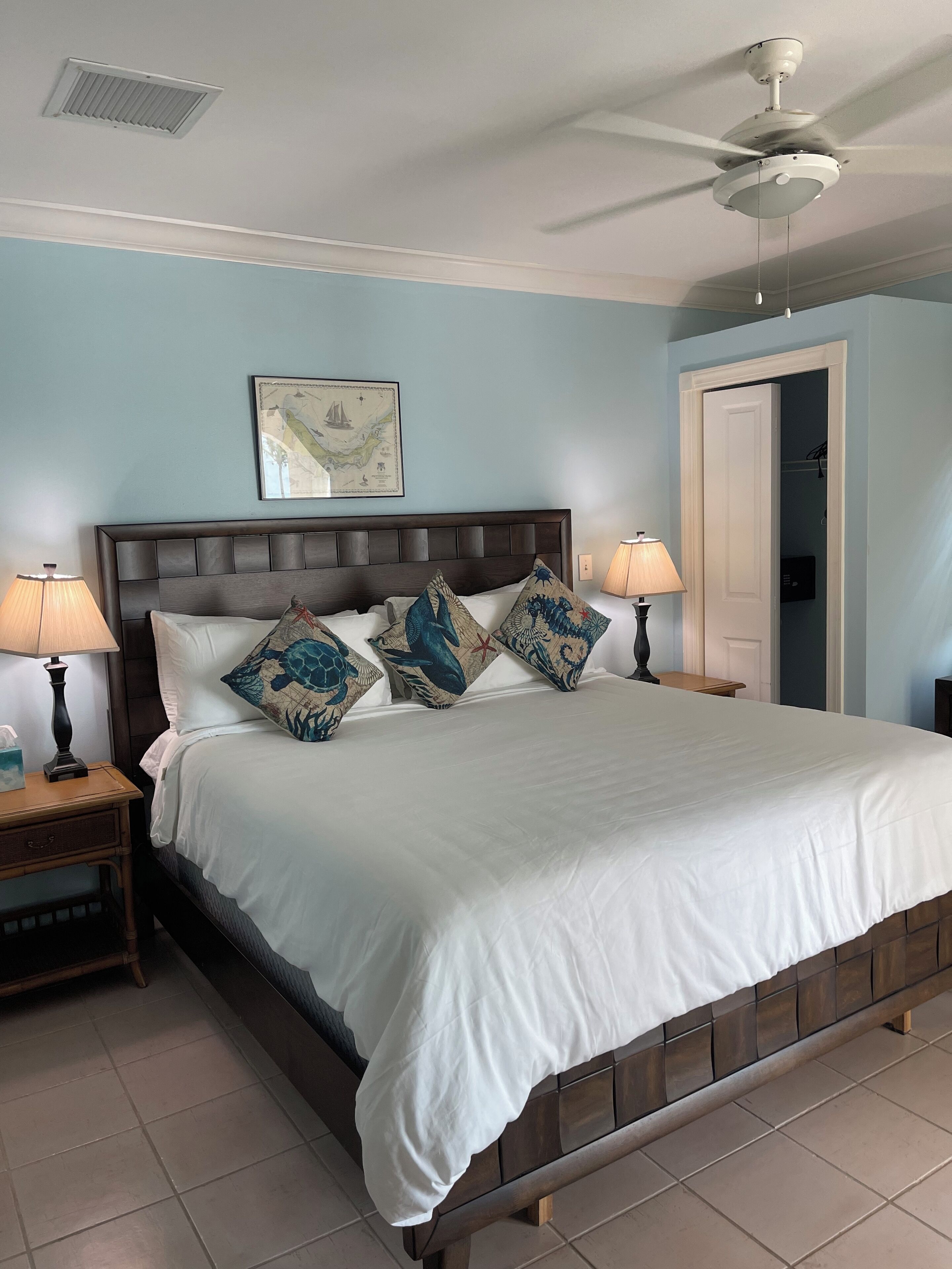 A bedroom with a large bed, two bedside tables, a ceiling fan, and a door leading to another room.