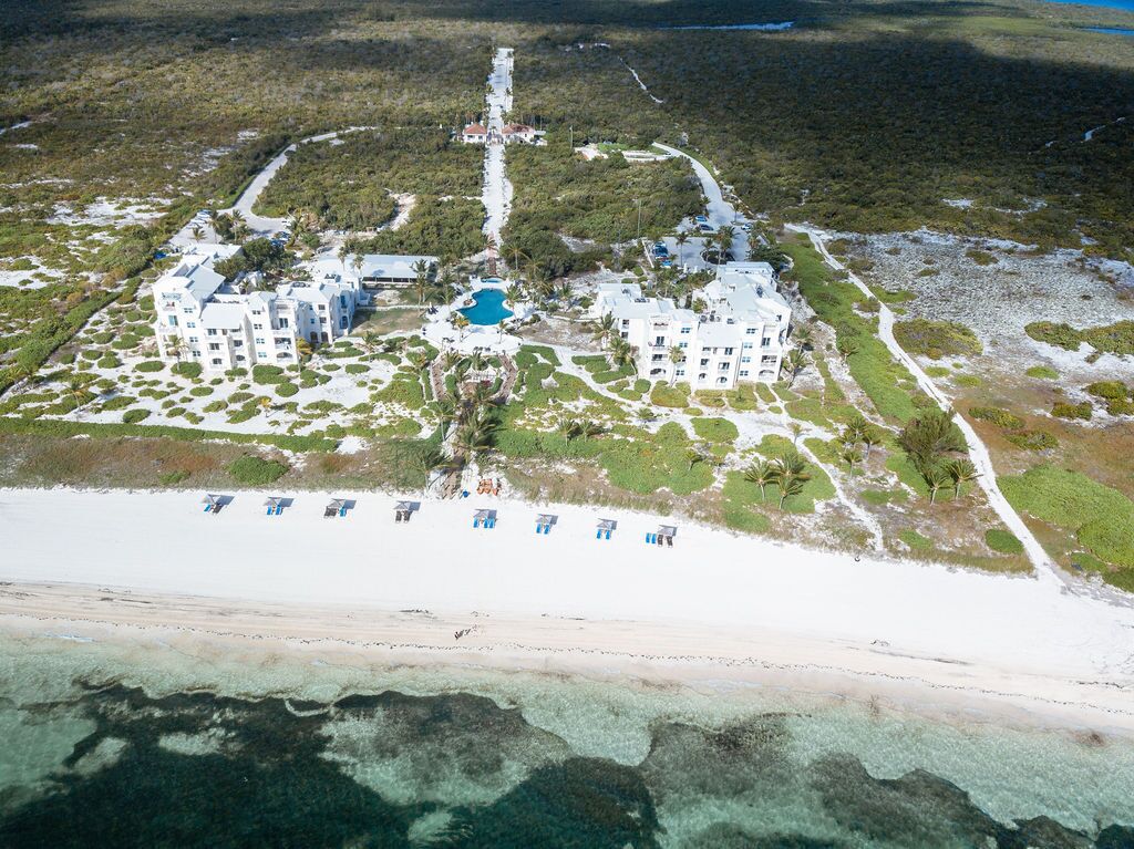 An aerial view of a resort with multiple buildings, a central pool, and a beach with umbrellas.