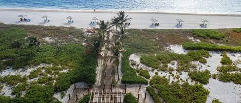 Vue aérienne d’un complexe hôtelier en bord de mer, avec une promenade en bois menant à une zone ombragée dotée de parasols et de chaises longues.