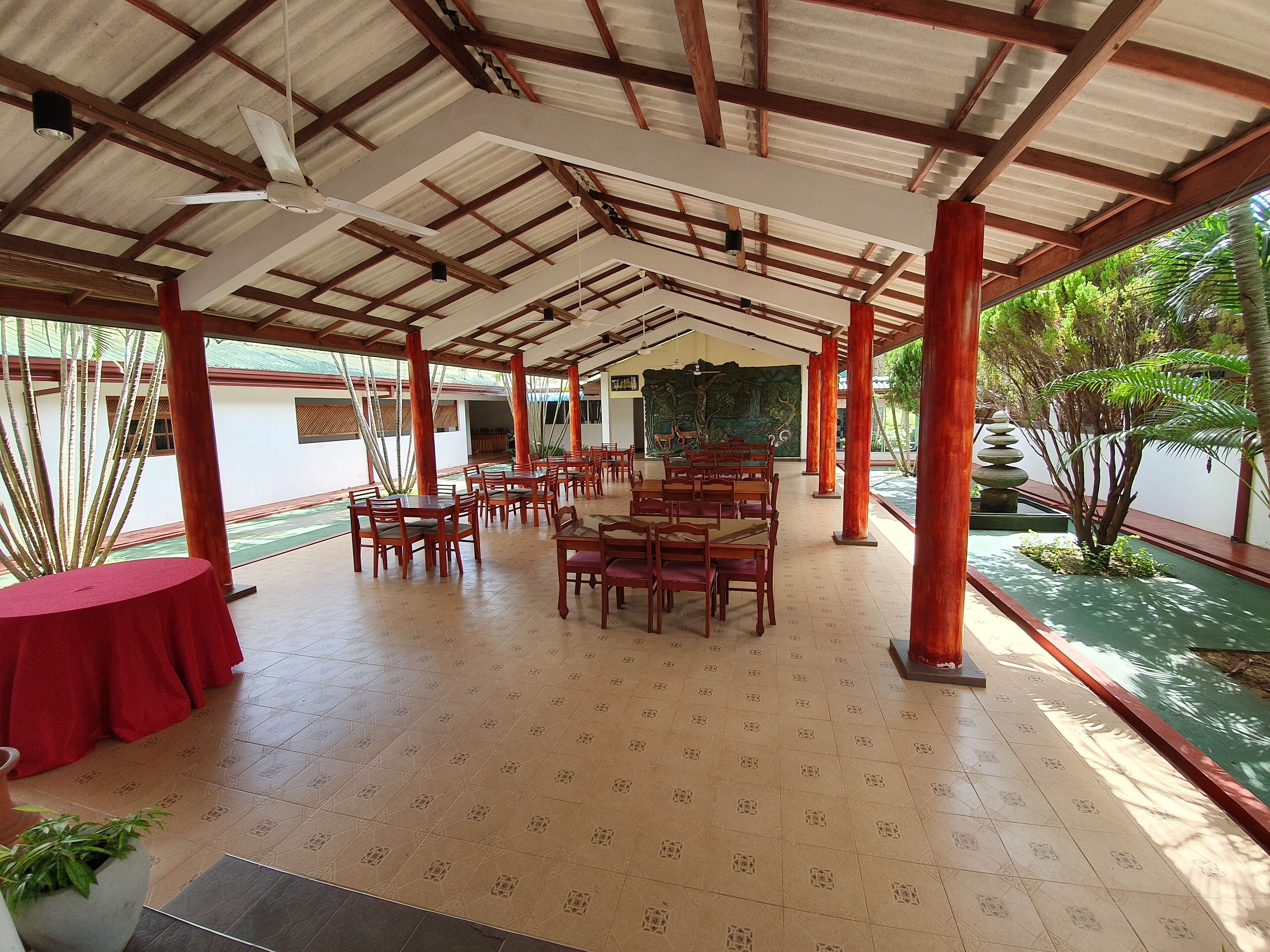 An indoor dining area with wooden tables and chairs, a tiled floor, and a ceiling with wooden beams and a fan.