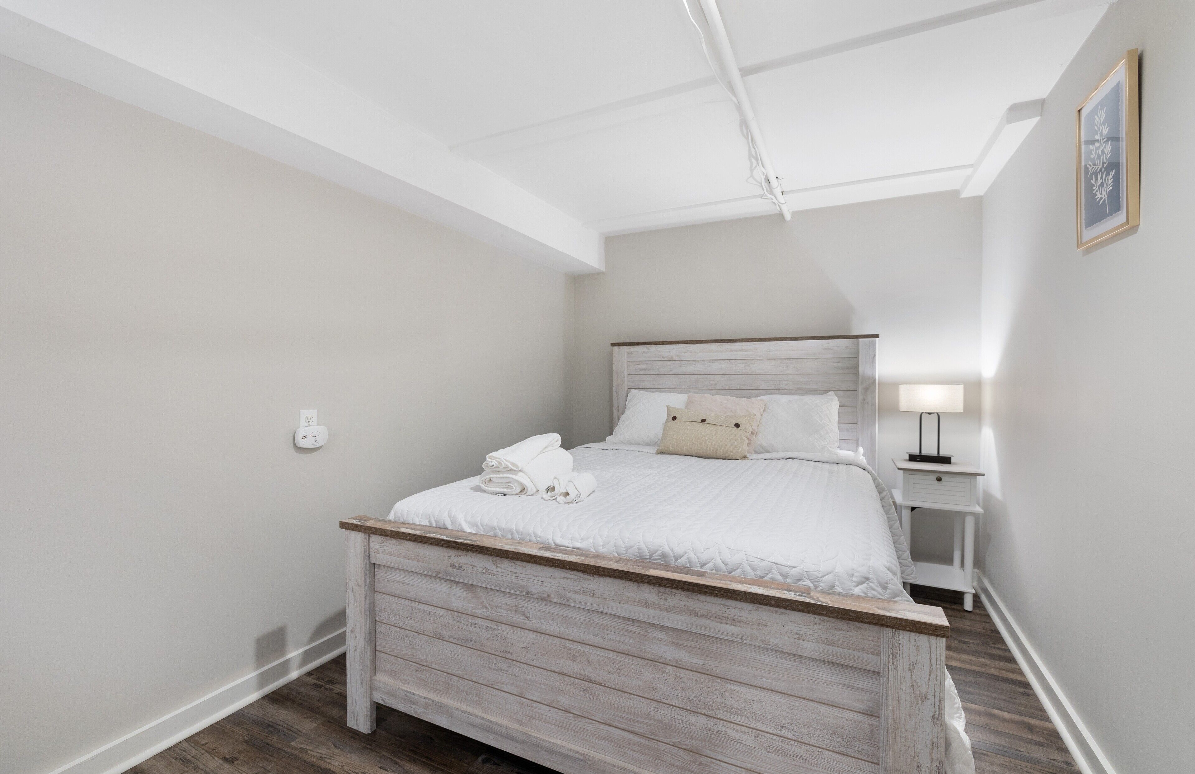 Un dormitorio con cama de madera, ropa de cama blanca, una mesita de noche con lámpara y un cuadro enmarcado en la pared.