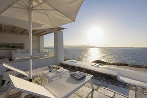 Un espace extérieur ensoleillé, aménagé avec des sièges, offrant une vue sur la mer, une piscine et un mur en pierre.