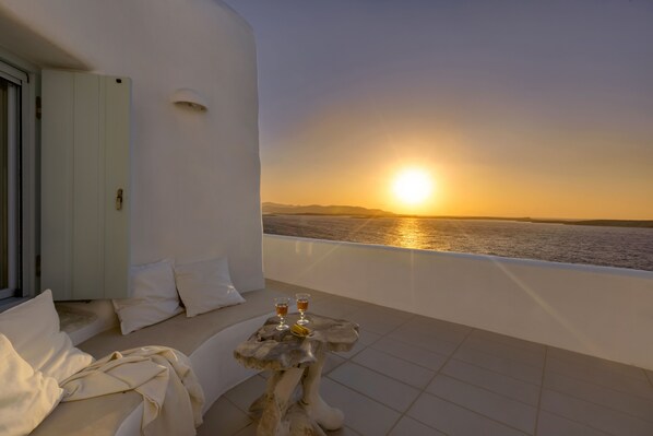 Ein Balkon mit Blick auf den Sonnenuntergang über dem Meer, ein Tisch mit zwei Gläsern und eine Couch mit Kissen.