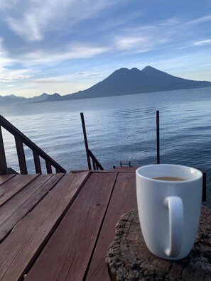 Une terrasse en bois avec une tasse blanche posée sur un dessous de verre, donnant sur une étendue d’eau calme et sur des montagnes lointaines.