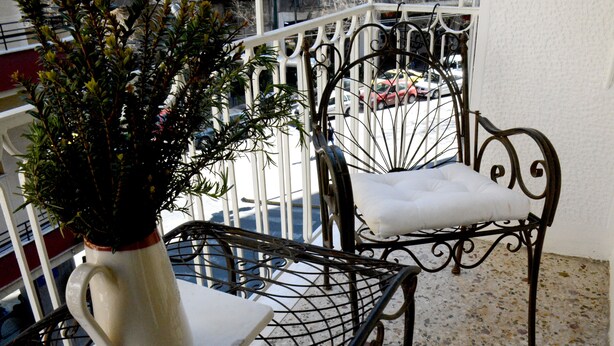 Un balcón con un jarrón blanco de plantas verdes, una silla de metal con cojín y una mesa de metal.