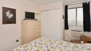Une chambre à coucher avec un lit, un téléviseur posé sur une commode, une fenêtre avec des stores et un appareil de climatisation.